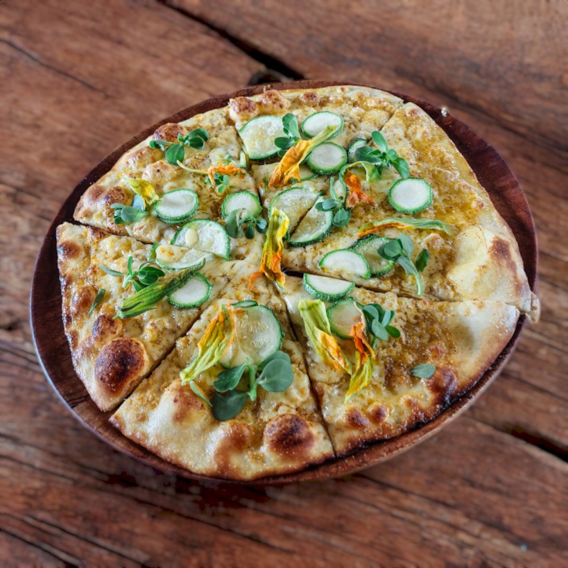 "Pizza gourmet con flores de calabaza, calabacín y microgreens sobre masa artesanal y queso derretido, servida en un plato rústico de madera."
